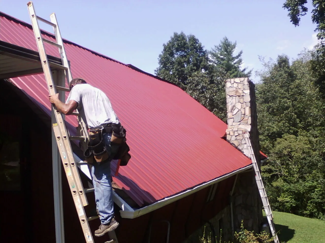 Roofer on a ladder beside a red metal roof and stone chimney — Soffit & Fascia Repair by Roofing Brothers in Belle Vernon